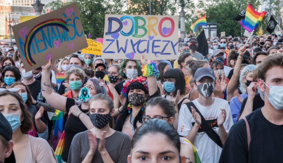 Demonstracja 8 sierpnia w Warszawie pod Pałacem Kultury i Nauki. W centralnej części kadru osoba trzyma wysoko kartonowy baner z napisem: "dobro zwycięży", w tęczowych kolorach.