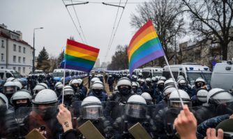 Protest, demonstacja, dwie małe tęczowe flagi trzymane w rękach tuż przed rzędem uzdrojonych policjantów.