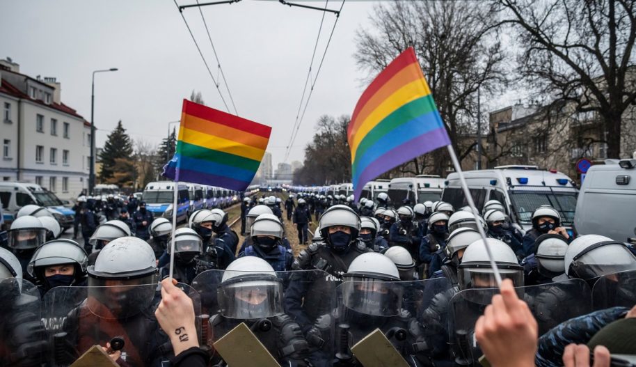 Protest, demonstacja, dwie małe tęczowe flagi trzymane w rękach tuż przed rzędem uzdrojonych policjantów.