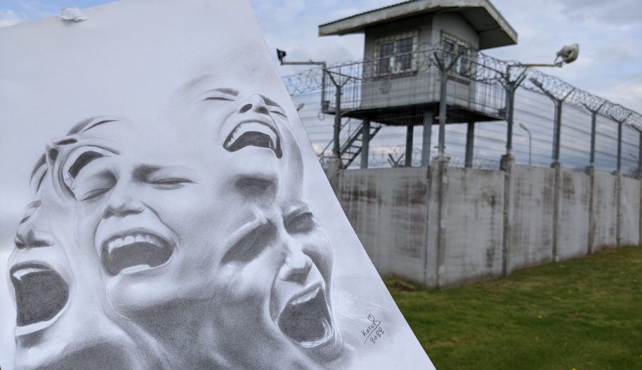 Zdjęcie przedstawia rysunek krzyczących postaci, a w tle wieża strażnicza, mury z drutem kolczastym - strzeżony ośrodek dla cudzoziemców