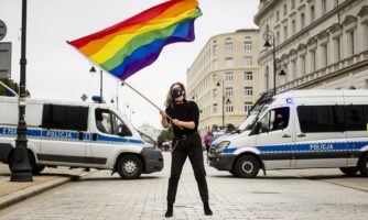 Protest przeciwko dyskryminacji osób LGBTI+ w Warszawie, 30 sierpnia 2020. Jedna z protestujących osób stoi pośrodku ulicy Krakowskie przedmieście, wymachując dużą tęczową flagą w rękach. Za nią stoją dwa radiowozy policyjne blokujące ulicę.