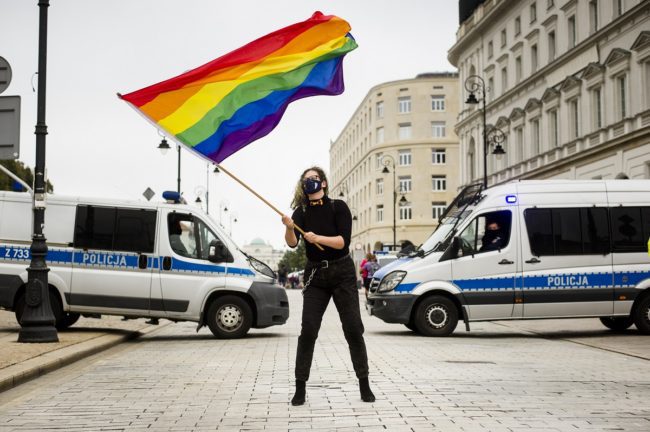 Protest przeciwko dyskryminacji osób LGBTI+ w Warszawie, 30 sierpnia 2020. Jedna z protestujących osób stoi pośrodku ulicy Krakowskie przedmieście, wymachując dużą tęczową flagą w rękach. Za nią stoją dwa radiowozy policyjne blokujące ulicę.