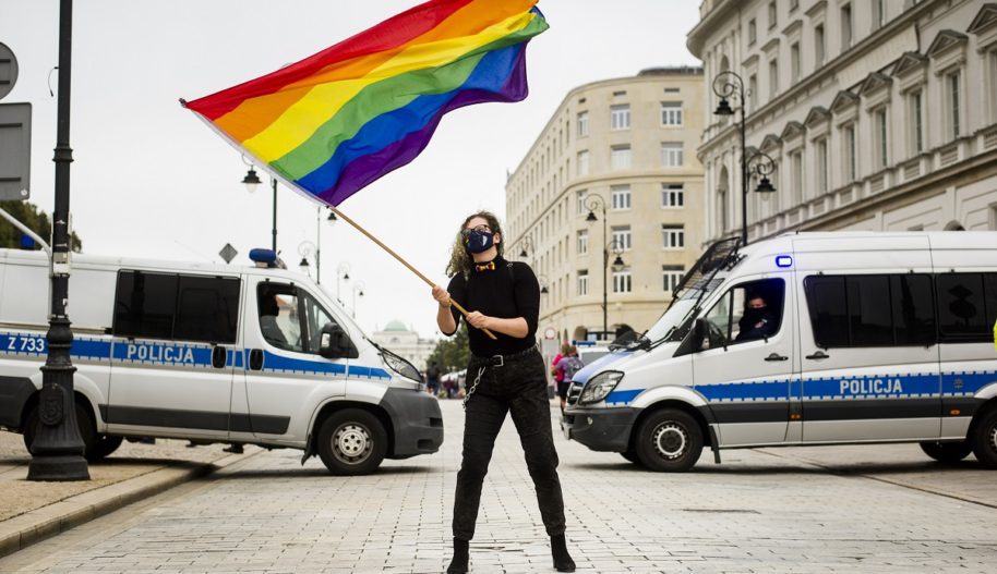 Protest przeciwko dyskryminacji osób LGBTI+ w Warszawie, 30 sierpnia 2020. Jedna z protestujących osób stoi pośrodku ulicy Krakowskie przedmieście, wymachując dużą tęczową flagą w rękach. Za nią stoją dwa radiowozy policyjne blokujące ulicę.
