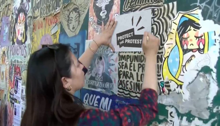 Stopklatka spotu do kampanii Protect the Protest. Aktywistka nalepia na śnianę wlepkę z logotypem kampanii Protect the Protest.