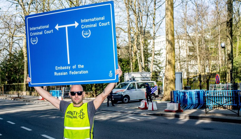 Demonstracja Amnesty International w Brukseli przed rosyjską ambasadą, 24 marca 2022. Aktywista w kamizelce z logiem Amnesty trzyma baner z drogowskazem pokazującym drogi z ambasady Rosji w kierunku Międzynarodowego Trybunału w Hadze.