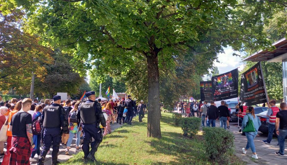 Dwie manifestacje idące ulicą, odgrodzone policjantami, w środku szpaler drzew.