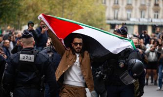 Mężczyzna trzymający flagę Palestyny podczas protestu w Paryżu, obok niego funkcjonariusze policji.