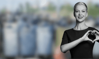 Uśmiechnięta kobieta pokazująca gest serca, w tle rozmazane grupy policji z tarczami.