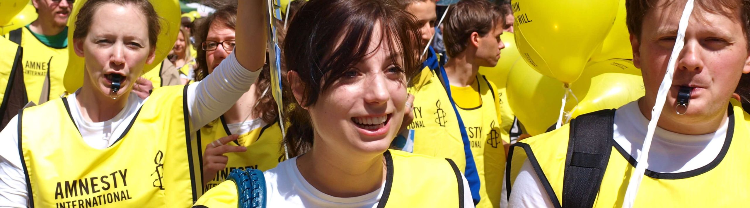 Młode osoby z bannerem Amnesty International na pokojowej manifestacji, dwie młode dziewczyny, trzymają balony, jedna dmucha w gwizdek