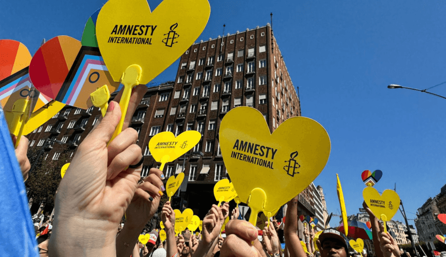 Rząd dłoni trzymających serduszka z logo Amnesty International, w tle duży budynek.