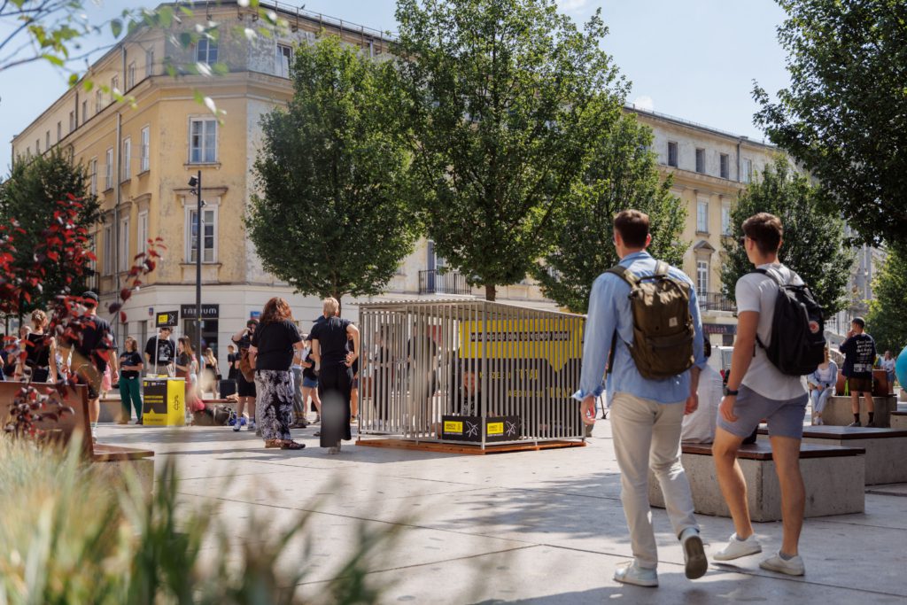 Symboliczna cela więzienna na placu. Obok drzewo i przechodzący ludzie.