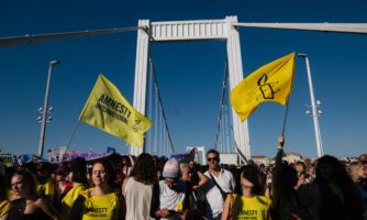 Marsz równości w Budapeszcie, na pierwszym planie osoby z flagami Amnesty. W tle filary mostu i czyste niebo.