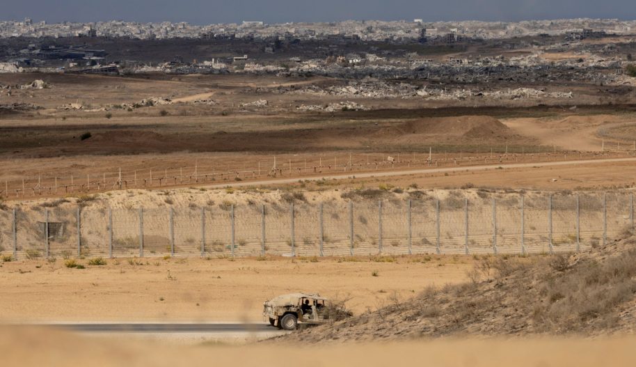 Pojazd wojskowy jadący wzdłuż zasieków, w tle zniszczone budynki w Gazie.