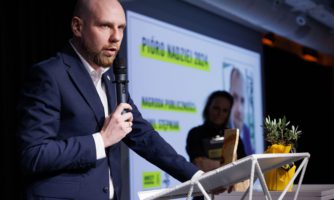 Mężczyzna mówiący do mikrofonu na scenie, w tle slajdy z prezentacją.