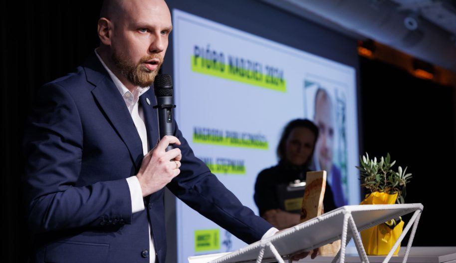 Mężczyzna mówiący do mikrofonu na scenie, w tle slajdy z prezentacją.