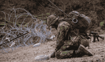 Klęczący żołnierz wyciąga z ziemi minę podczas ćwiczeń wojskowych, w tle druty kolczaste i las.