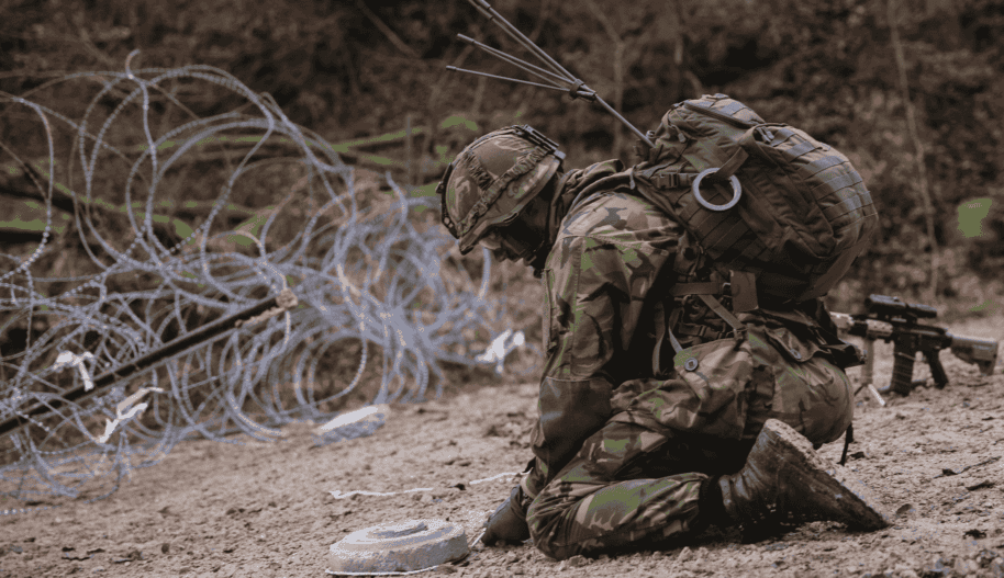 Klęczący żołnierz wyciąga z ziemi minę podczas ćwiczeń wojskowych, w tle druty kolczaste i las.