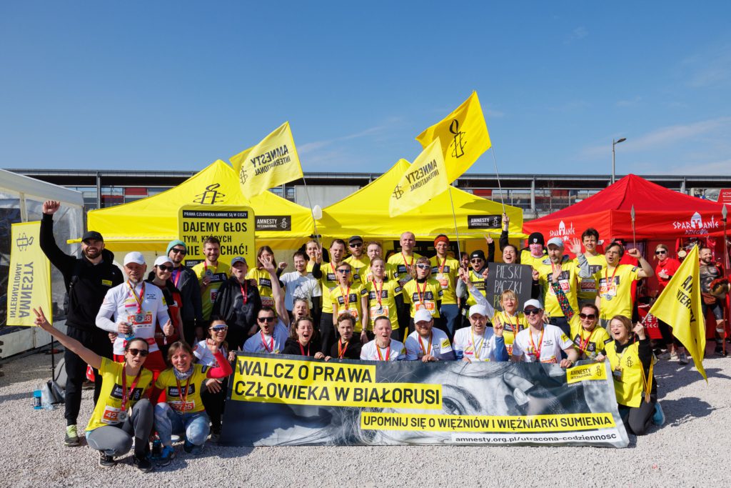 Duża grupa osób pozuje do zdjęcia. Z przodu baner przypominający o białoruskich więźniach i więźniarkach politycznych, w tle namiot z logotypami Amnesty.