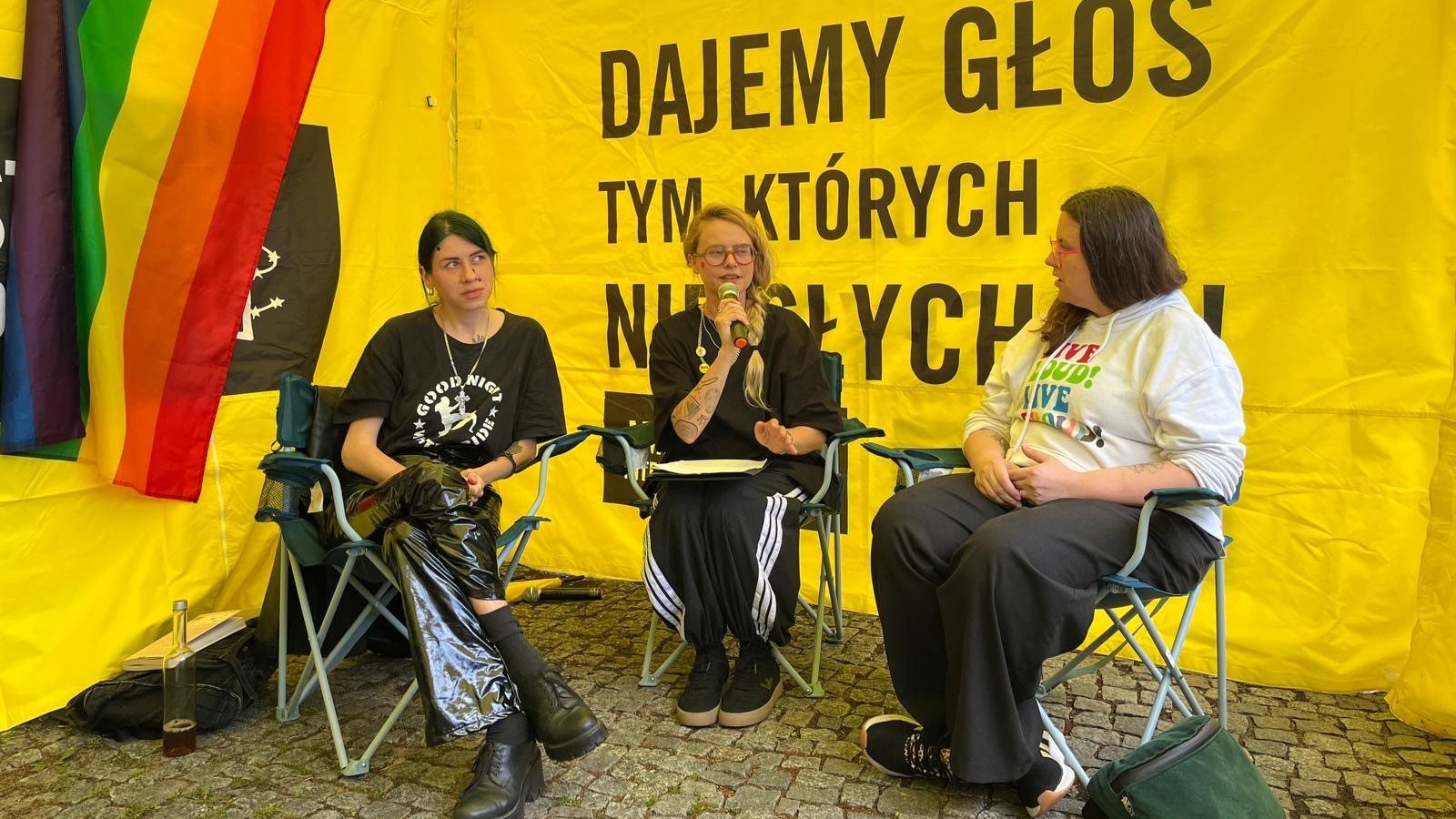 Trzy osoby siedzące w dużym namiocie w trakcie debaty, w tle symbole społeczności LGBTI i hasła Amnesty International.